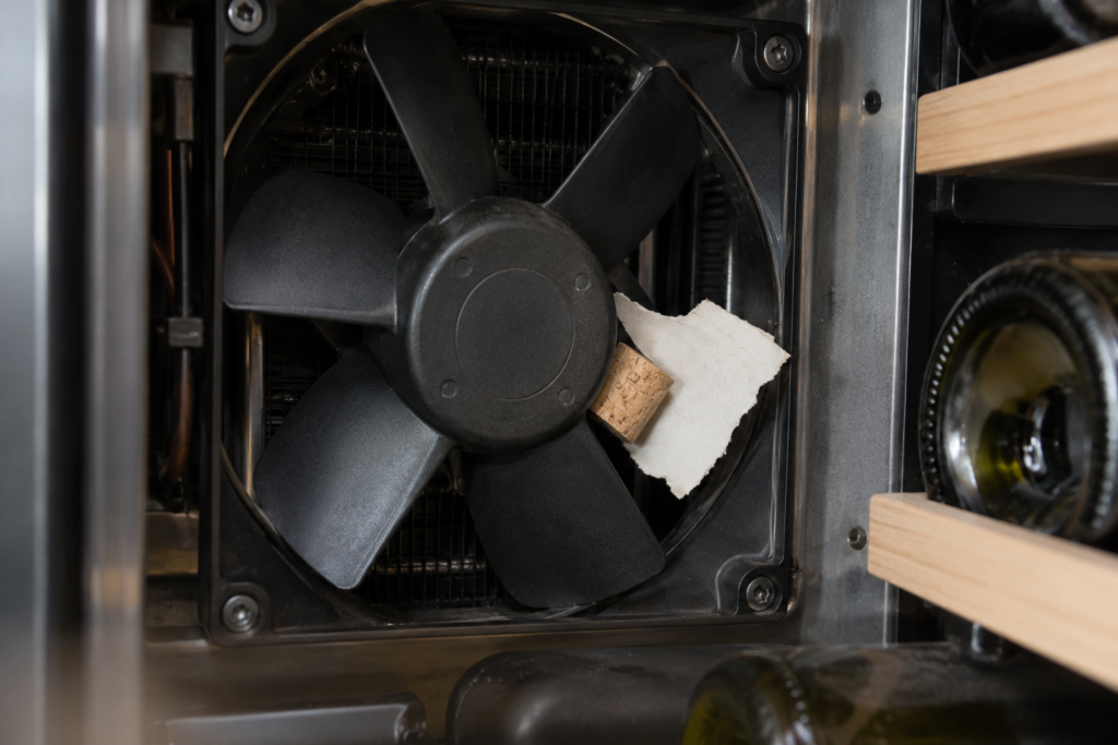 A fan blade inside a wine cabinet with a torn bottle label and a cork fragment wedged against its edge, close view with soft light.